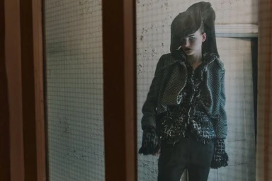 Model wearing a dark textured fringed tweed jacket and sculptural hood against a rustic interior wall.
