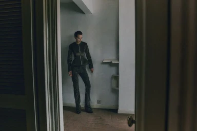 Model standing in an interior room wearing a dark structured jacket and matching trousers shot through a doorway