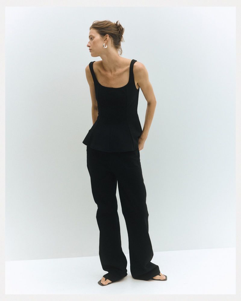 A fair-skinned adult woman poses in a black top and loose black pants against a white backdrop in a studio setting, looking left.