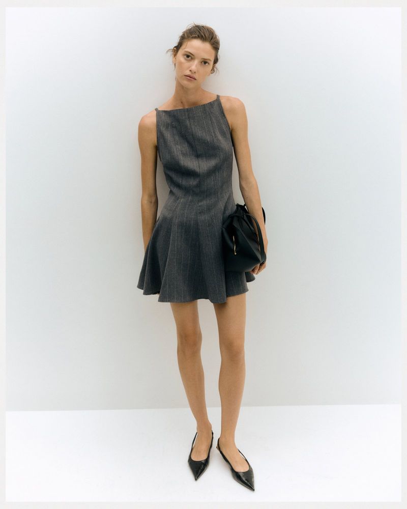 A slender woman poses in a gray pinstripe mini dress with a flared skirt, accessorized with black flats and a small black handbag.