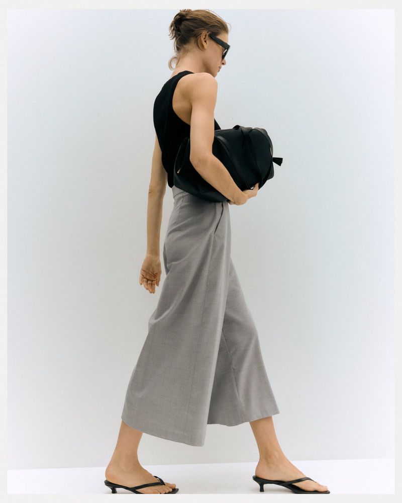 Model Valerie Scherzinger in profile walking in a black tank top, wide-leg grey culottes, and black sandals against a white background.