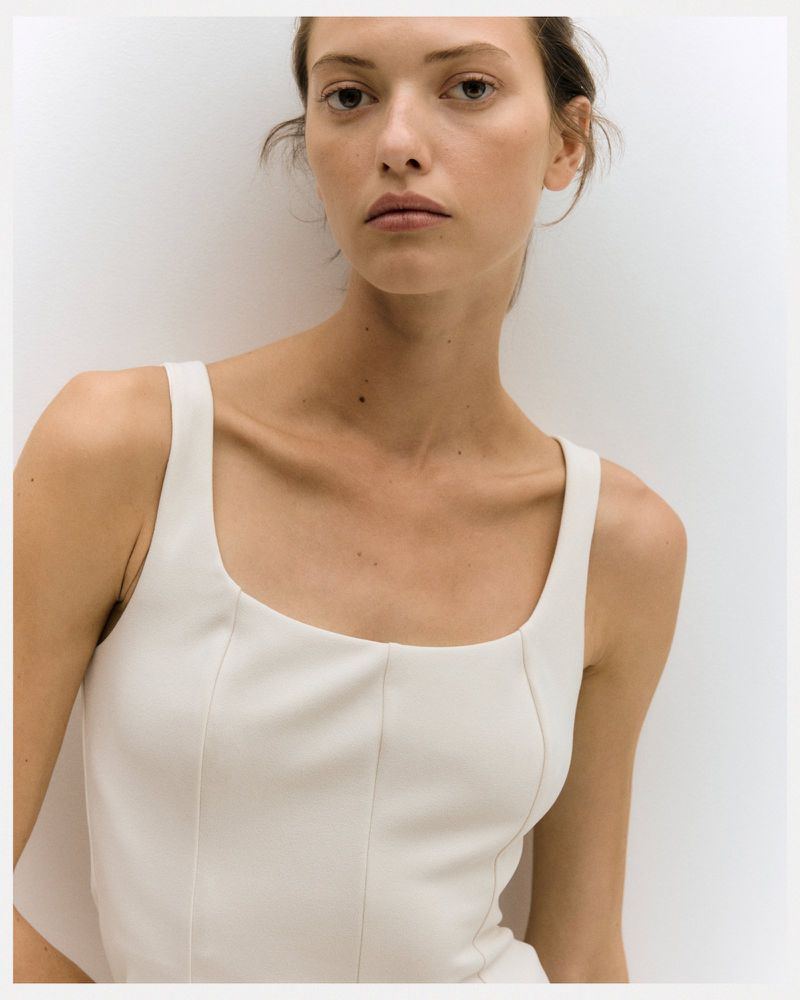 Close-up of a woman with neutral expression wearing a white top, posing against a light background in a portrait style fashion shot.