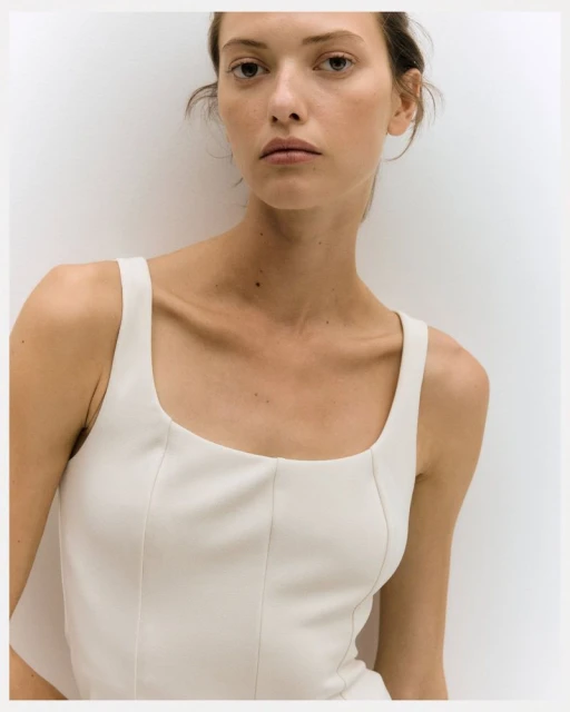 Close-up of a woman with neutral expression wearing a white top, posing against a light background in a portrait style fashion shot.