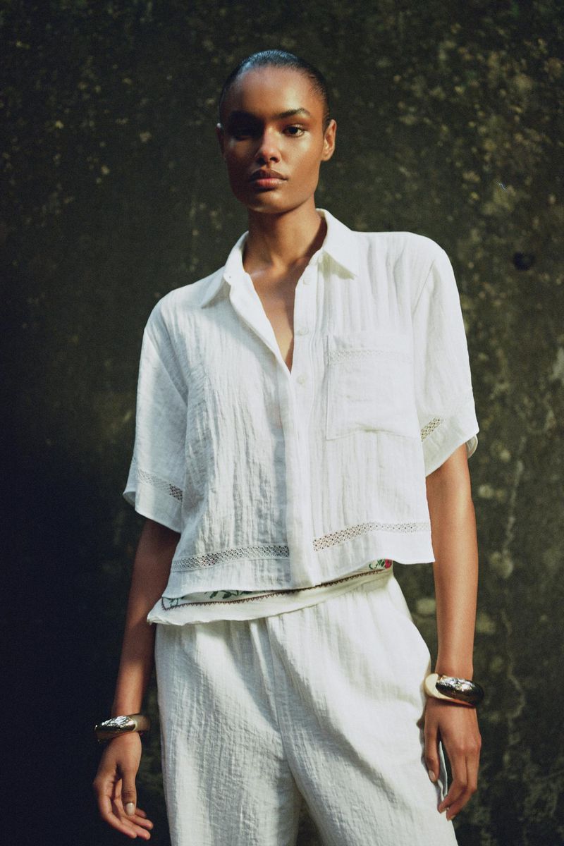 Elegant woman in a white shirt and pants posing in front of a textured green wall, showcasing a minimalist fashion aesthetic.