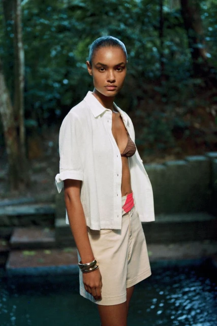 An adult woman in a white button-down shirt and khaki shorts stands outdoors with trees and dark water in the background, posing for fashion content.