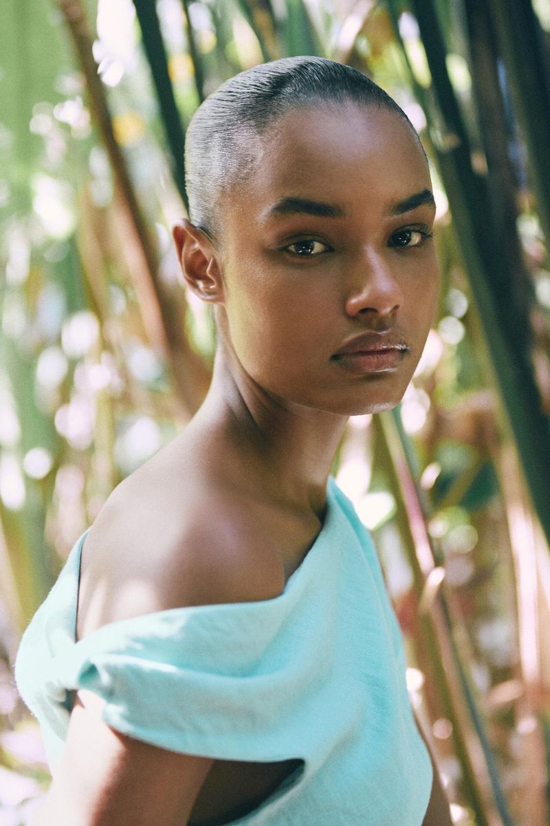 Portrait of a Black woman in a turquoise off-the-shoulder dress, posing outdoors with blurred green foliage in the background.
