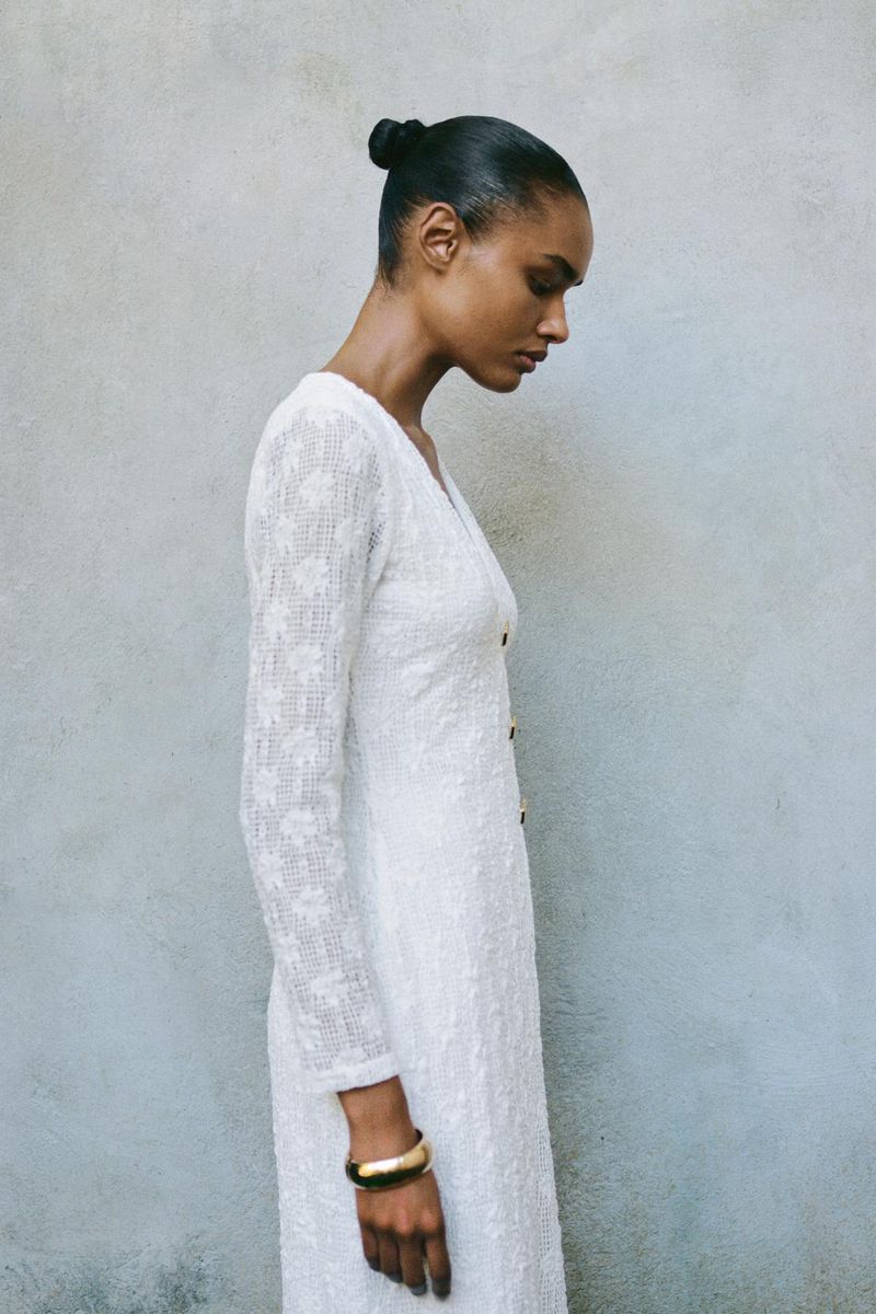 Side profile shot of a Black female adult in a long sleeve white dress and a gold bracelet against a textured grey background.