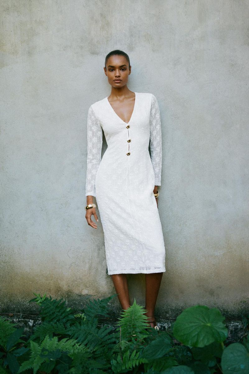 Elegant adult female model in long-sleeve buttoned dress posing in front of a concrete wall with green foliage at the bottom.