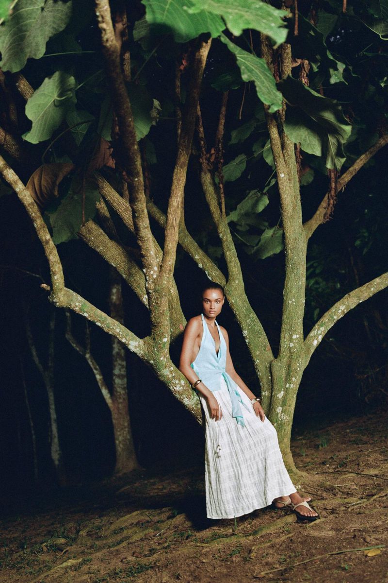 Woman in long white skirt and light blue top leans against a tree in an outdoor setting, gazing directly at the camera.