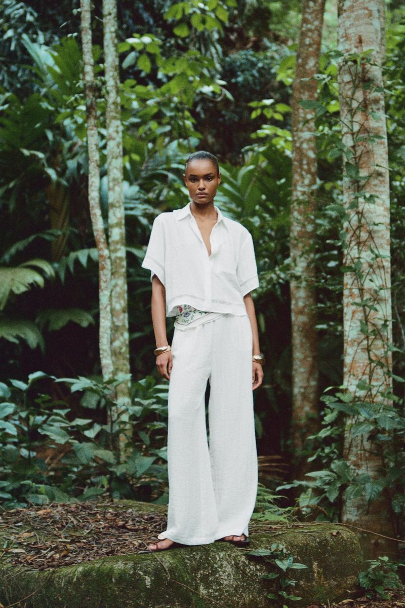 A woman poses in white shirt and wide-leg pants on a rock, surrounded by a lush forest, creating a fashion editorial shot.