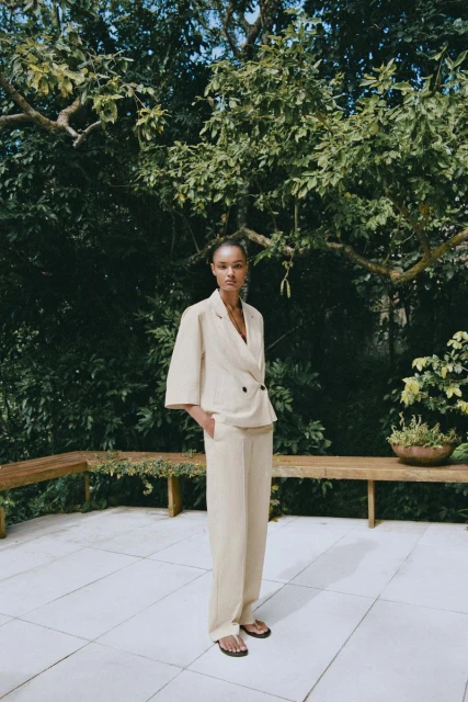Adult female model in a light beige suit stands on a tiled patio with benches, surrounded by green foliage and trees in daylight.