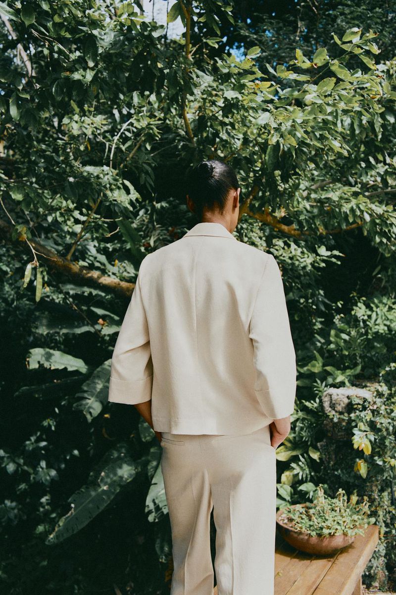 Back view of an adult in a light beige suit outdoors surrounded by green foliage, creating a natural, muted-tone fashion image.