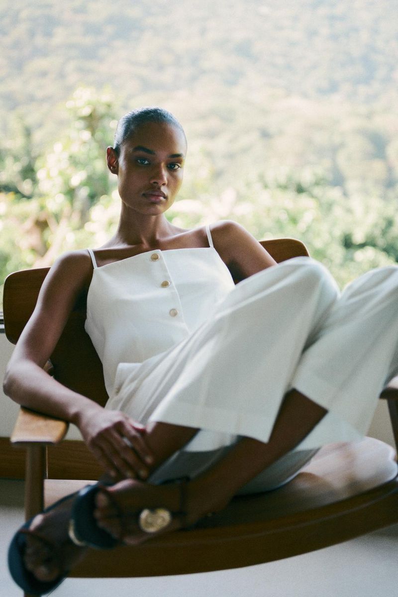 Woman in a white linen two-piece outfit reclines on a wooden rocking chair with an outdoor foliage background in soft, natural light.