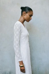 Ana Beatriz Cortes wearing a long white lace dress and gold cuff bracelet in profile against a textured grey wall.