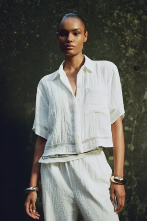 Model wearing a white short-sleeve button-down shirt and trousers against a dark textured background for Zara A/W25