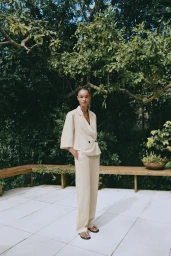 Model Ana Beatriz Cortes in a beige short-sleeve pantsuit standing on a stone terrace backed by lush greenery for Zara A/W25.