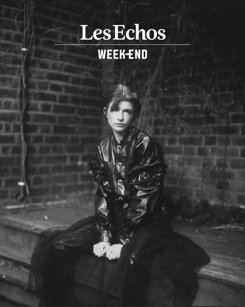 Black and white portrait of a young adult woman in a black jacket and skirt, seated against a brick wall, fashion editorial.