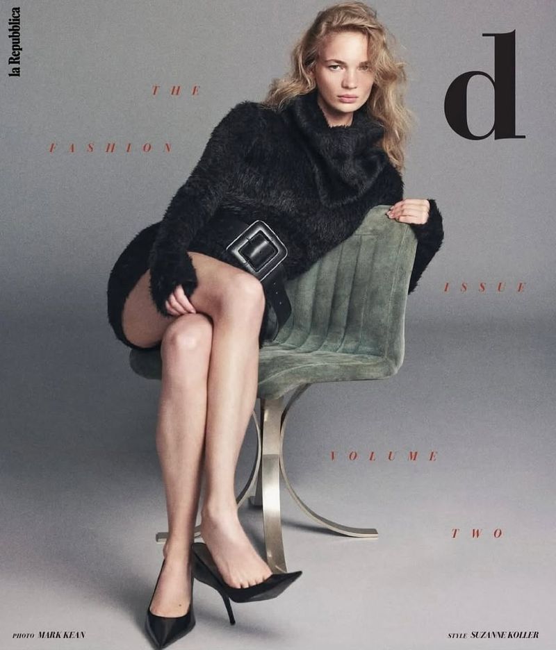 Fashion magazine cover featuring a woman in a black sweater and skirt sitting on an olive green chair in a grey studio setting.