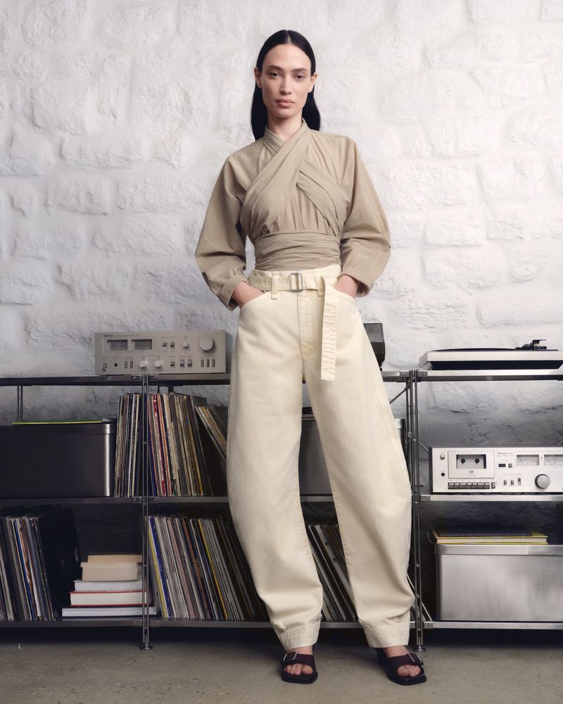 A fashionable adult female stands in a studio wearing a khaki wrap top, baggy denim pants, and dark sandals in a fashion editorial shot.