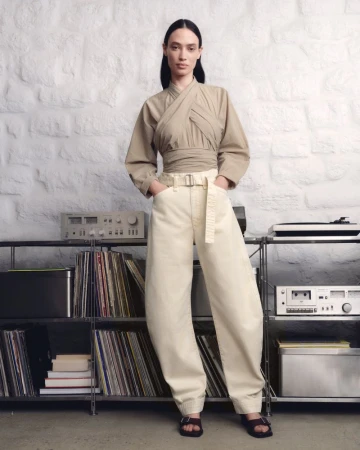A fashionable adult female stands in a studio wearing a khaki wrap top, baggy denim pants, and dark sandals in a fashion editorial shot.