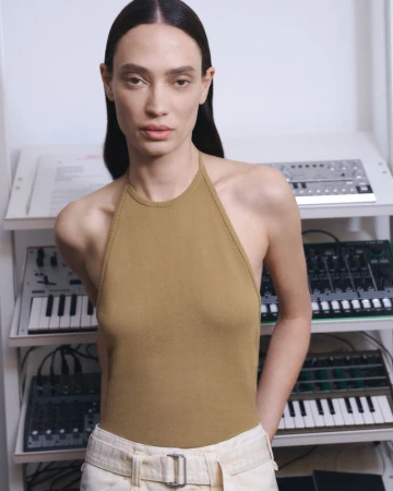 A brunette woman with neutral expression wears a tan halter top and cream trousers, posing indoors in front of sound equipment.