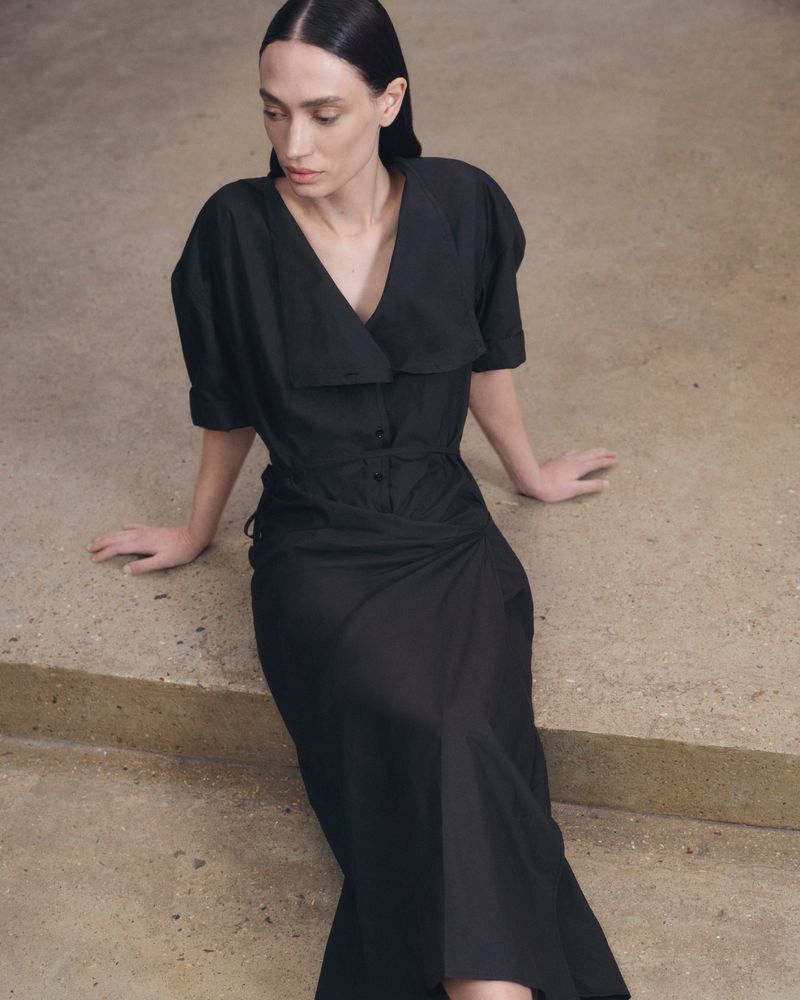 A slender woman in a black dress sits on concrete stairs, her dark hair neatly styled, creating a striking fashion editorial image.