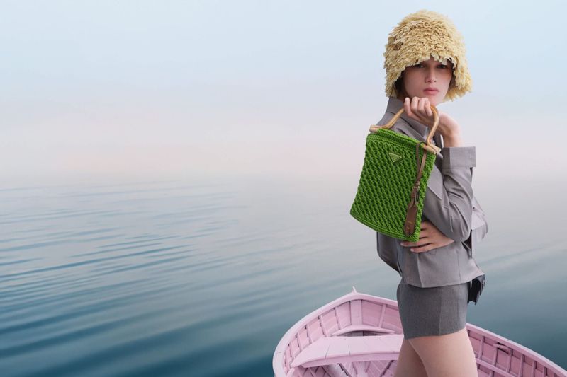 A stylish woman in boat holds a green woven Prada purse, wearing a bucket hat and grey outfit, against a serene blue ocean backdrop.