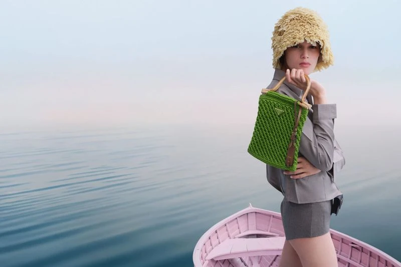 A stylish woman in boat holds a green woven Prada purse, wearing a bucket hat and grey outfit, against a serene blue ocean backdrop.