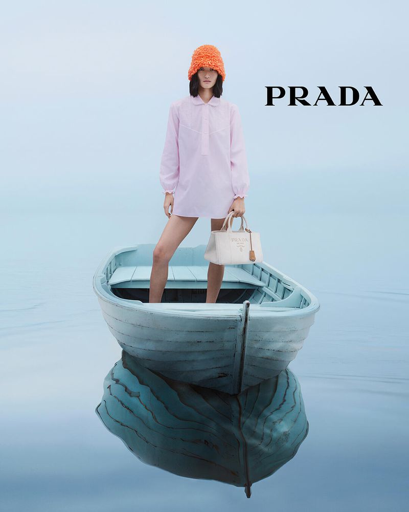 Fashion shot of a model standing in a blue rowboat on water, holding a white Prada purse, wearing a pink dress and an orange hat.