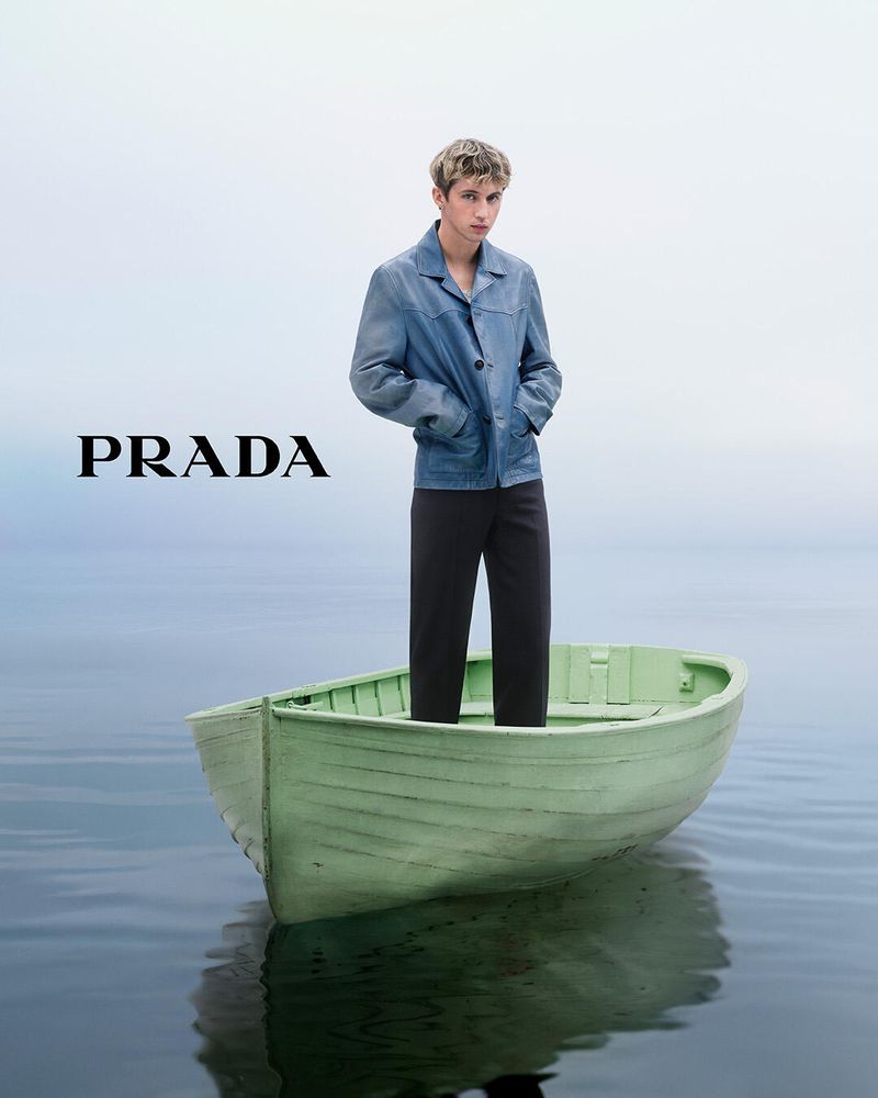 A fair-skinned young man stands in a green rowboat on calm water, wearing a denim jacket and black pants, with the Prada logo visible.