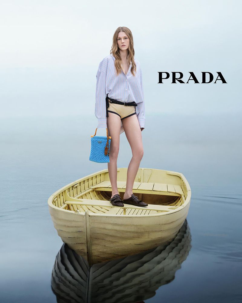 A fair-skinned fashion model poses in a yellow rowboat on the water with a blue woven handbag, wearing a blue striped shirt and neutral shorts.