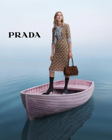 A woman stands in a pink boat on calm water, wearing a patterned dress, heels, and carrying a fringed handbag, with the Prada logo visible.