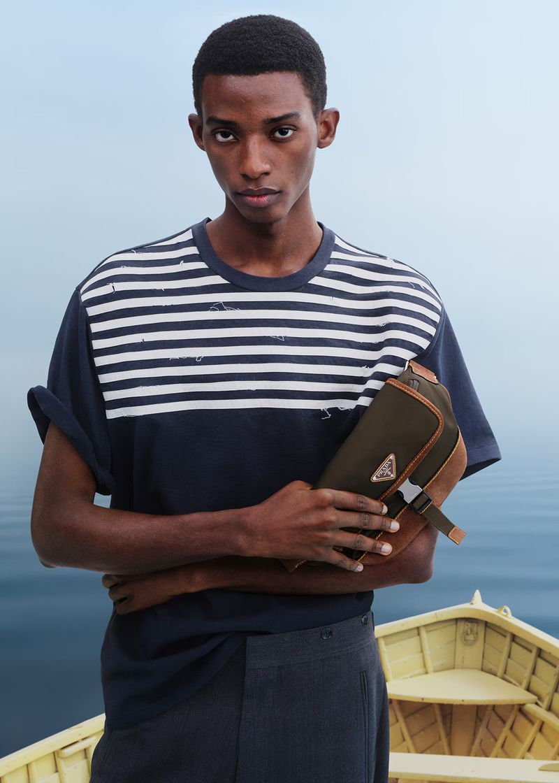 Dark-skinned male model in striped shirt holds a Prada bag on a boat with a light blue sky and calm ocean in the background.