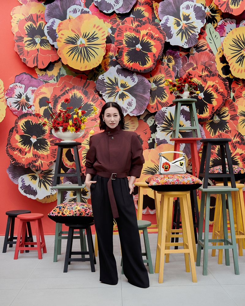 A woman poses in front of a vibrant floral display and colorful stools, wearing a brown shirt and black pants, with a patterned purse.