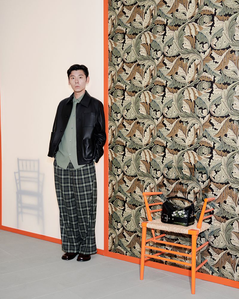A full shot of a man in a black jacket and plaid pants standing indoors next to a chair and patterned wallpaper, styled fashion photography.