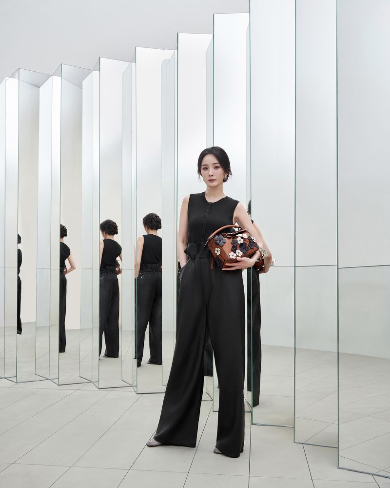 Elegant woman in black sleeveless top and wide-leg pants poses in a minimalist setting with mirrors, holding a floral purse.