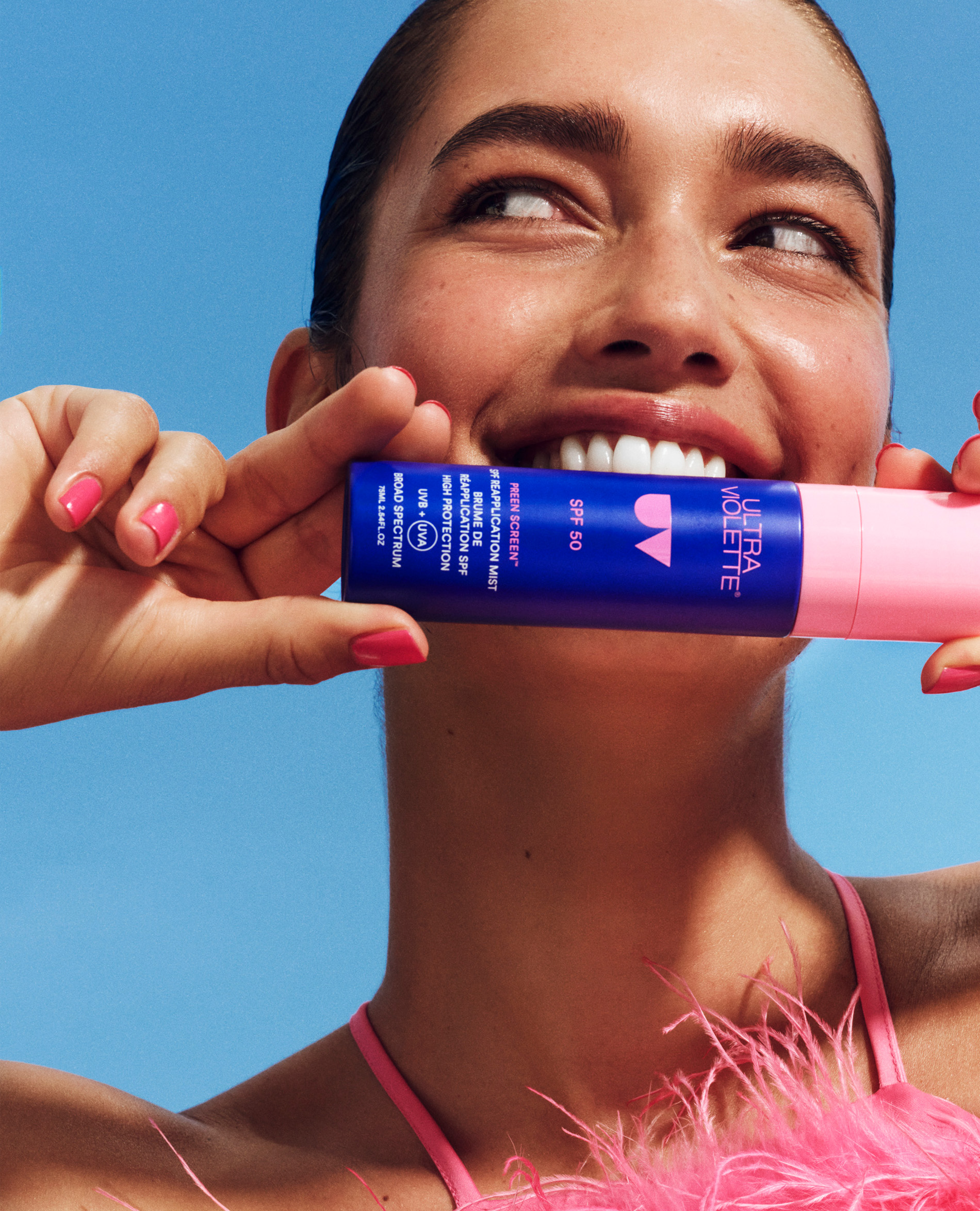 Smiling woman with pink nails holds a blue Ultra Violette sunscreen tube against a bright blue sky, showcasing beauty and sun protection.
