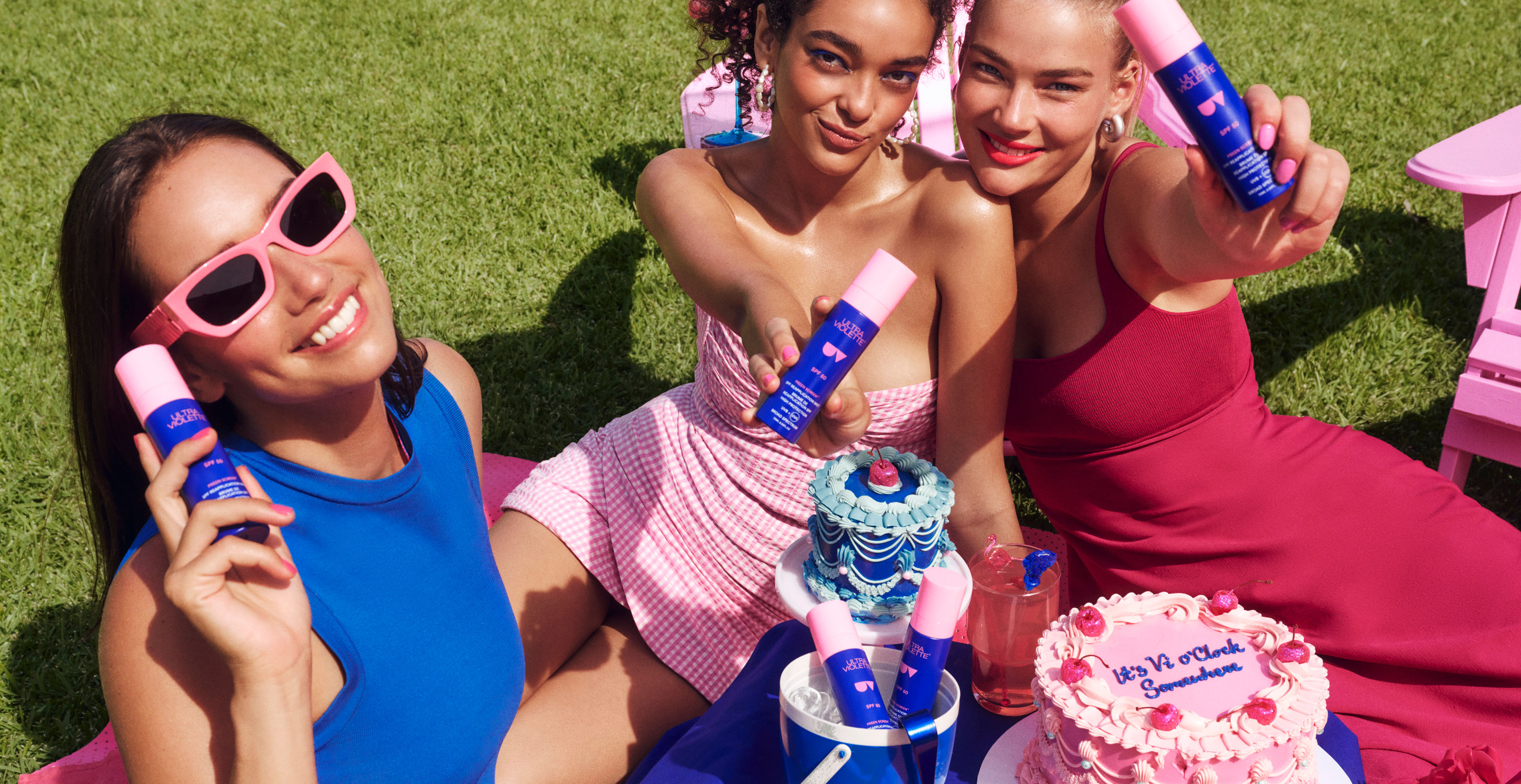 Three young adult women smile and hold SPF sunscreen on a blanket with cakes in outdoor setting, bathed in natural light, sunny day.