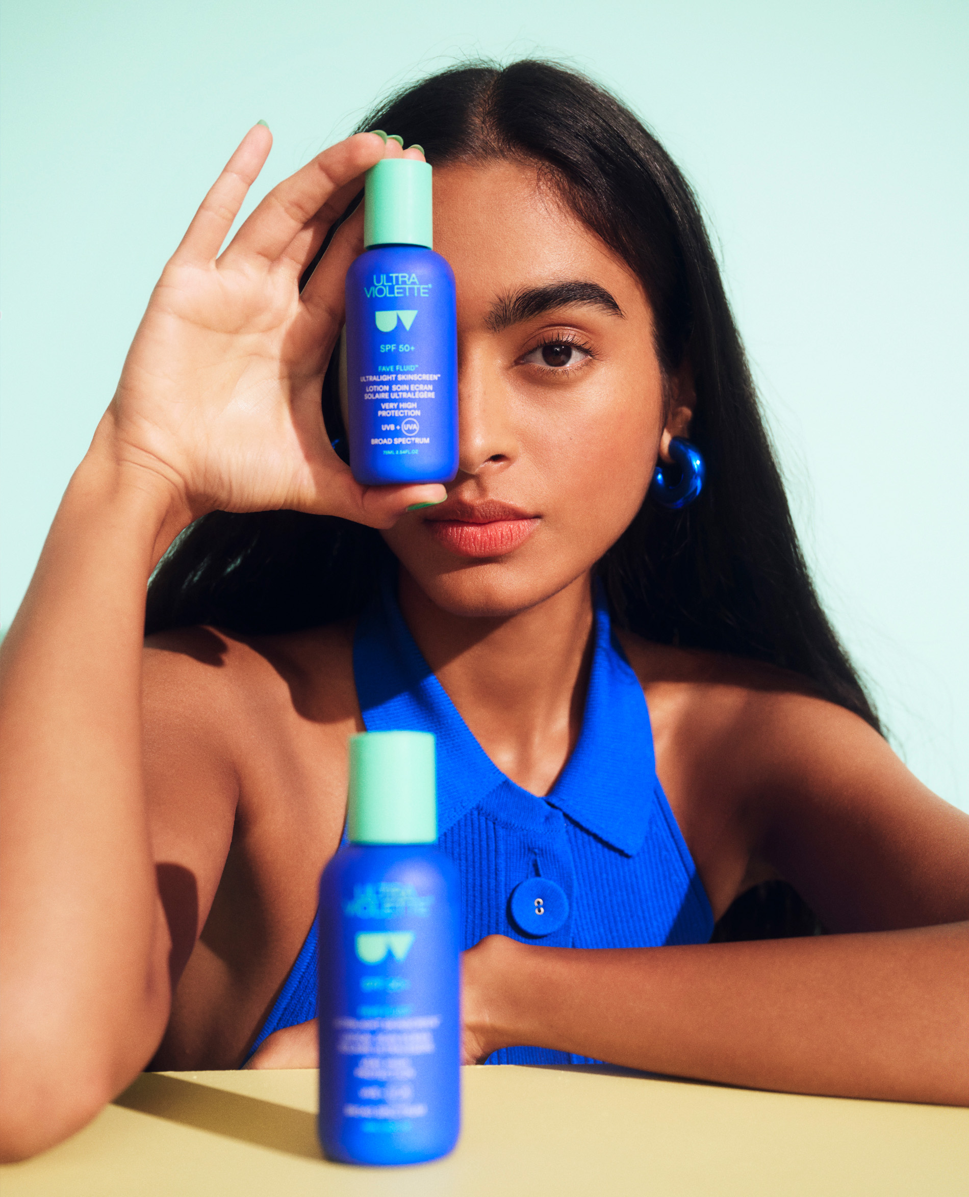 Woman poses with blue SPF bottle, light turquoise backdrop, bright light, yellow table. Beauty, skincare, UV protection concept.