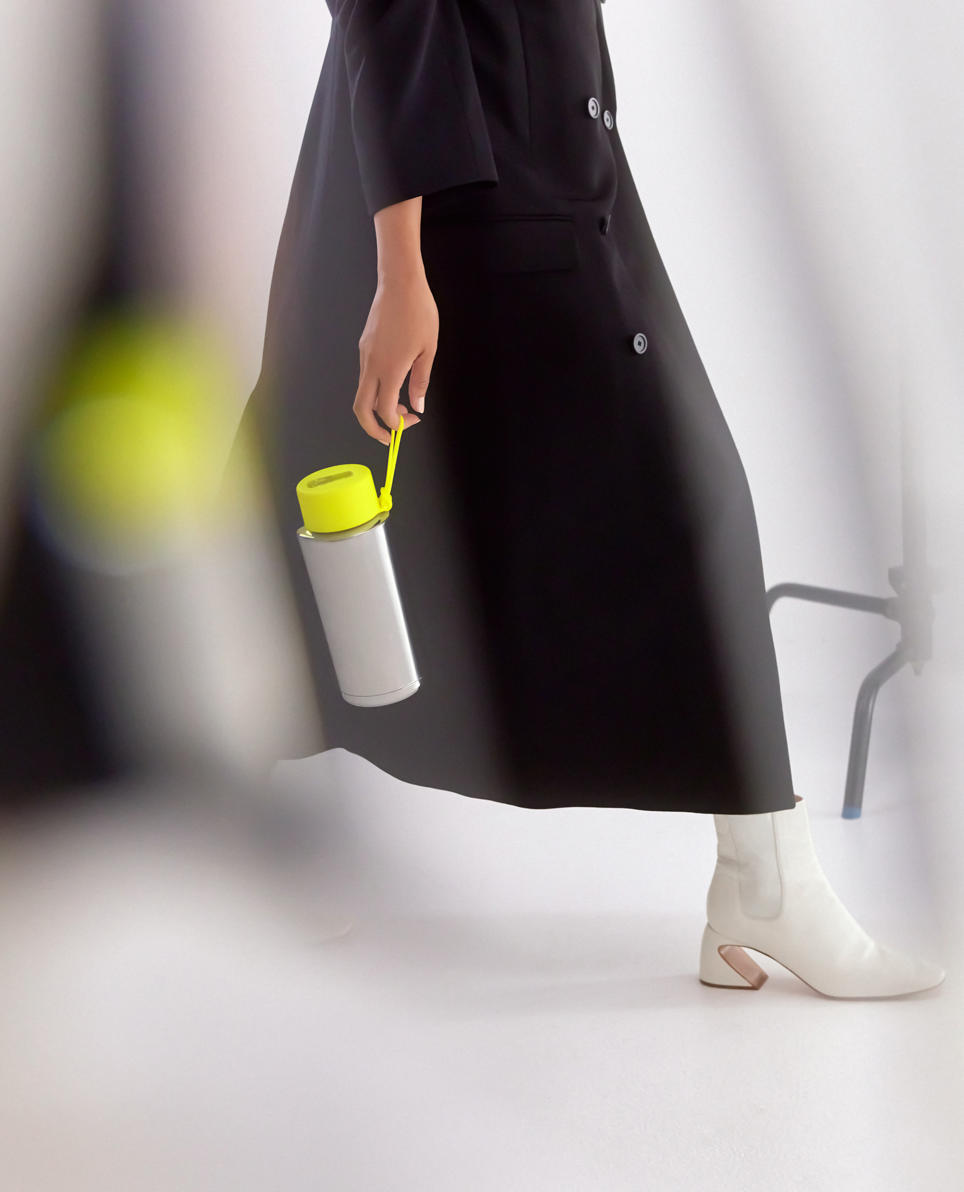 An adult female model carries a grey water bottle with a neon-yellow cap in a studio, wearing a black coat and white boots.