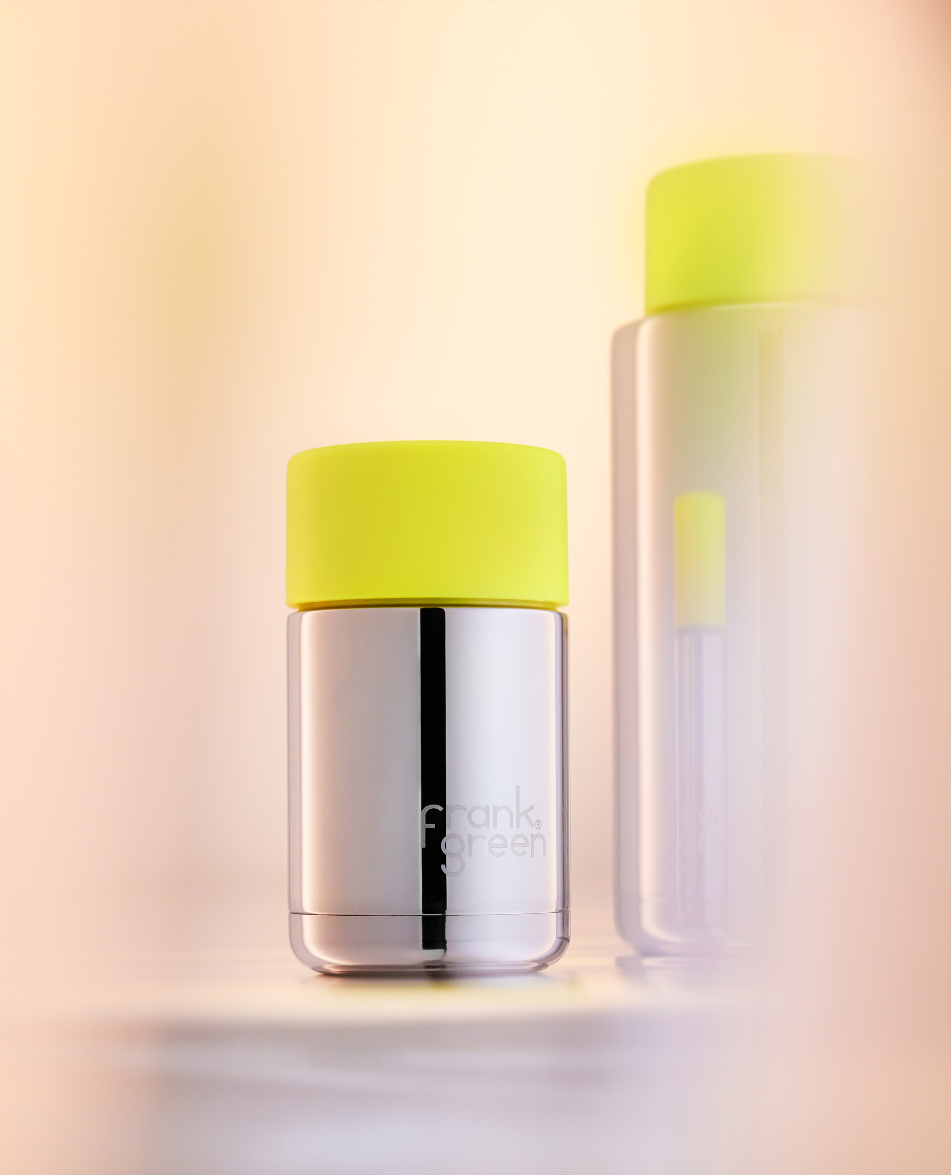 Two Frank Green water bottles on a neutral surface, one in focus with a stainless steel body and a neon yellow lid.