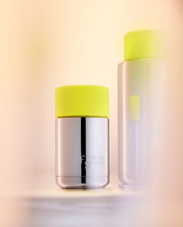Two Frank Green water bottles on a neutral surface, one in focus with a stainless steel body and a neon yellow lid.