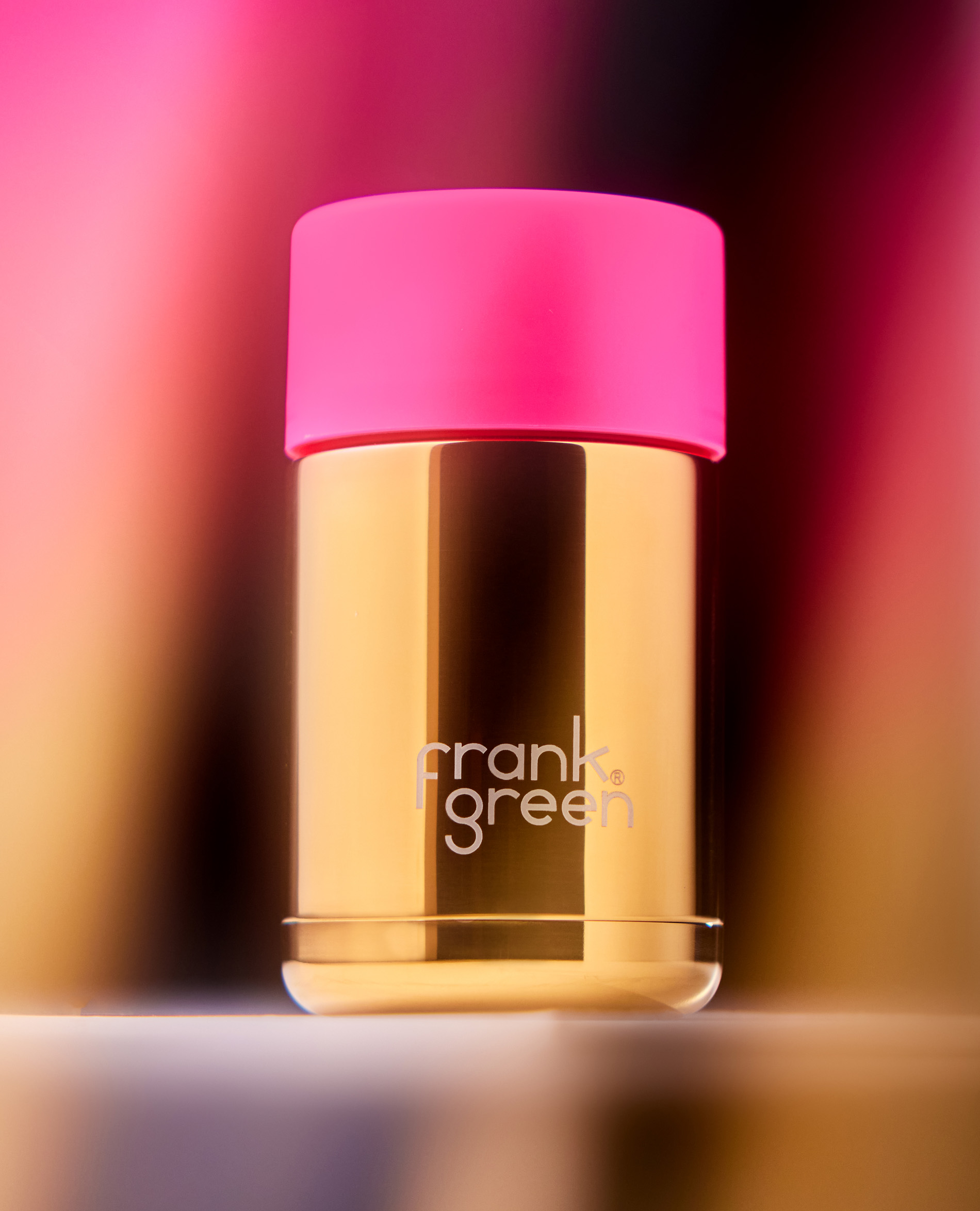Close-up studio shot of a gold Frank Green bottle with a pink lid against a soft-focus gradient background. Modern design.