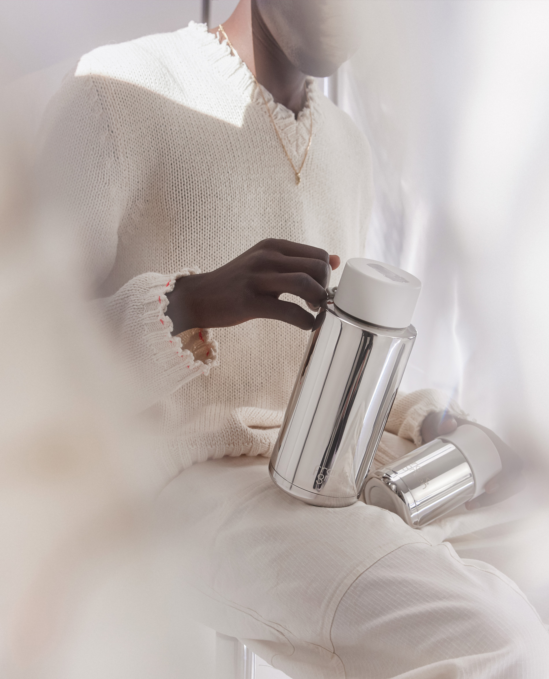 Person in white sweater holding a silver insulated water bottle in a softly lit, neutral indoor setting with window light.