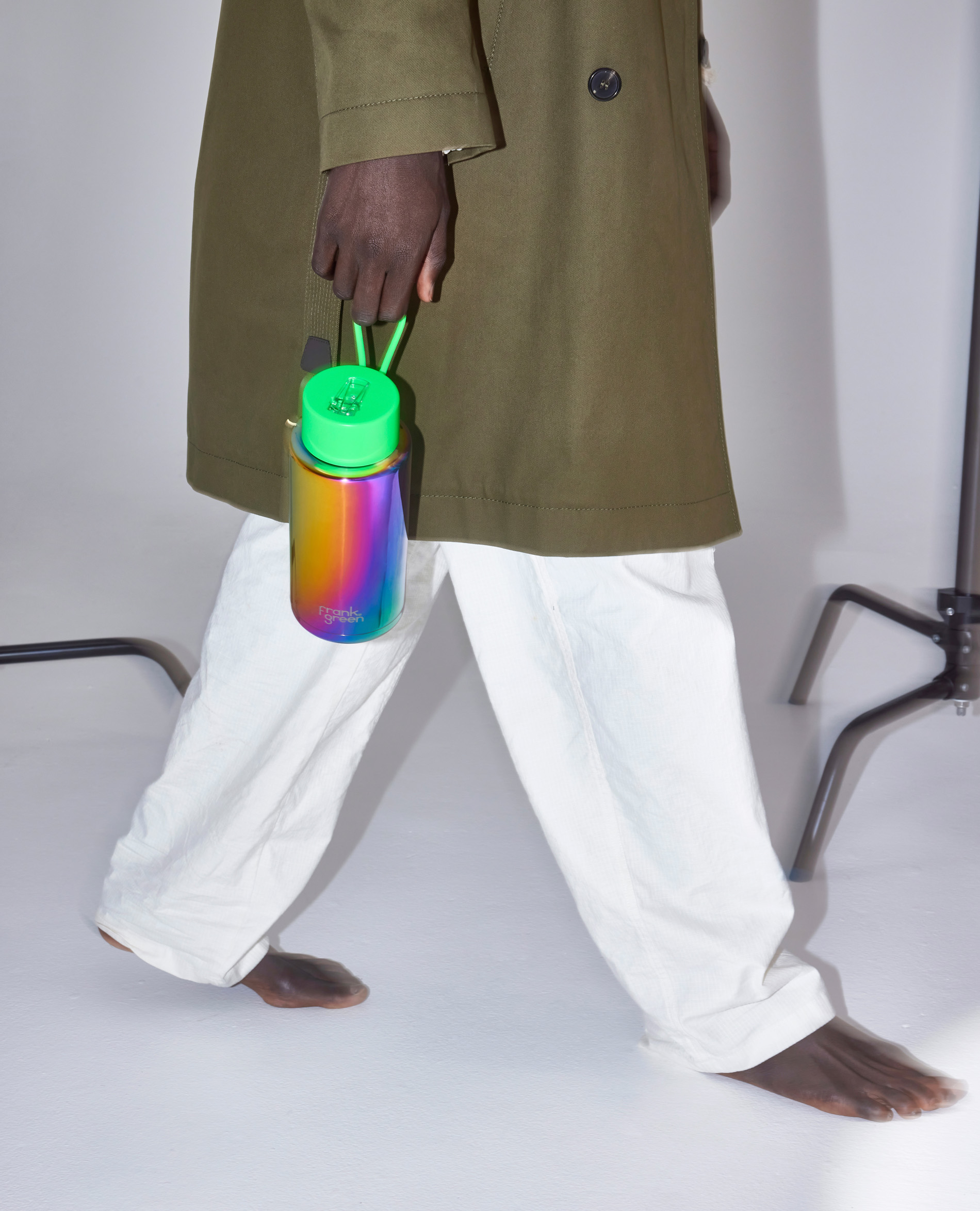Barefoot model wearing a trench coat and white pants holds a rainbow-colored water bottle in a studio with a plain background.