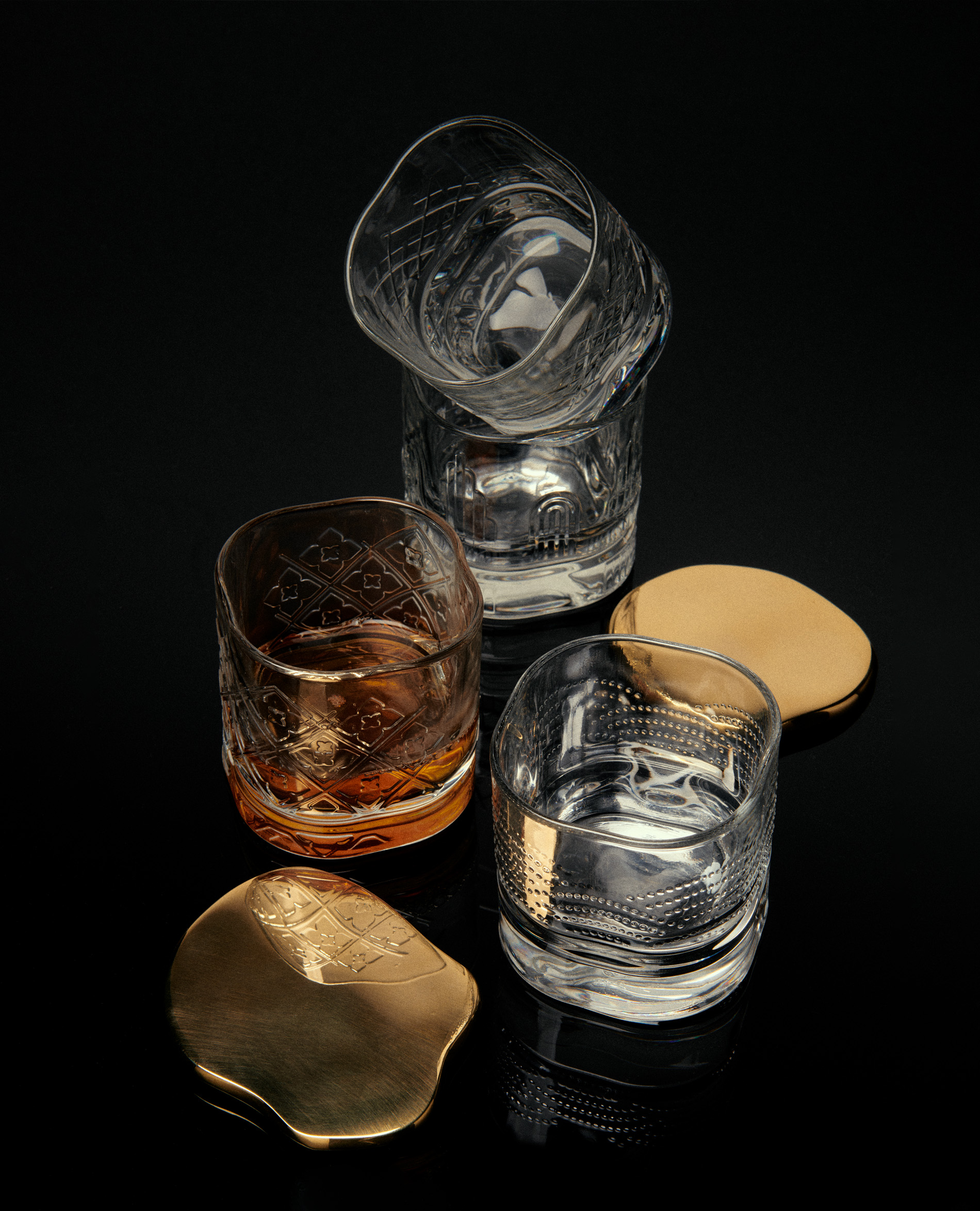 Elevated product shot showcases an arrangement of crystal glasses and gold coasters on a stark black background, highlighting textures and details.