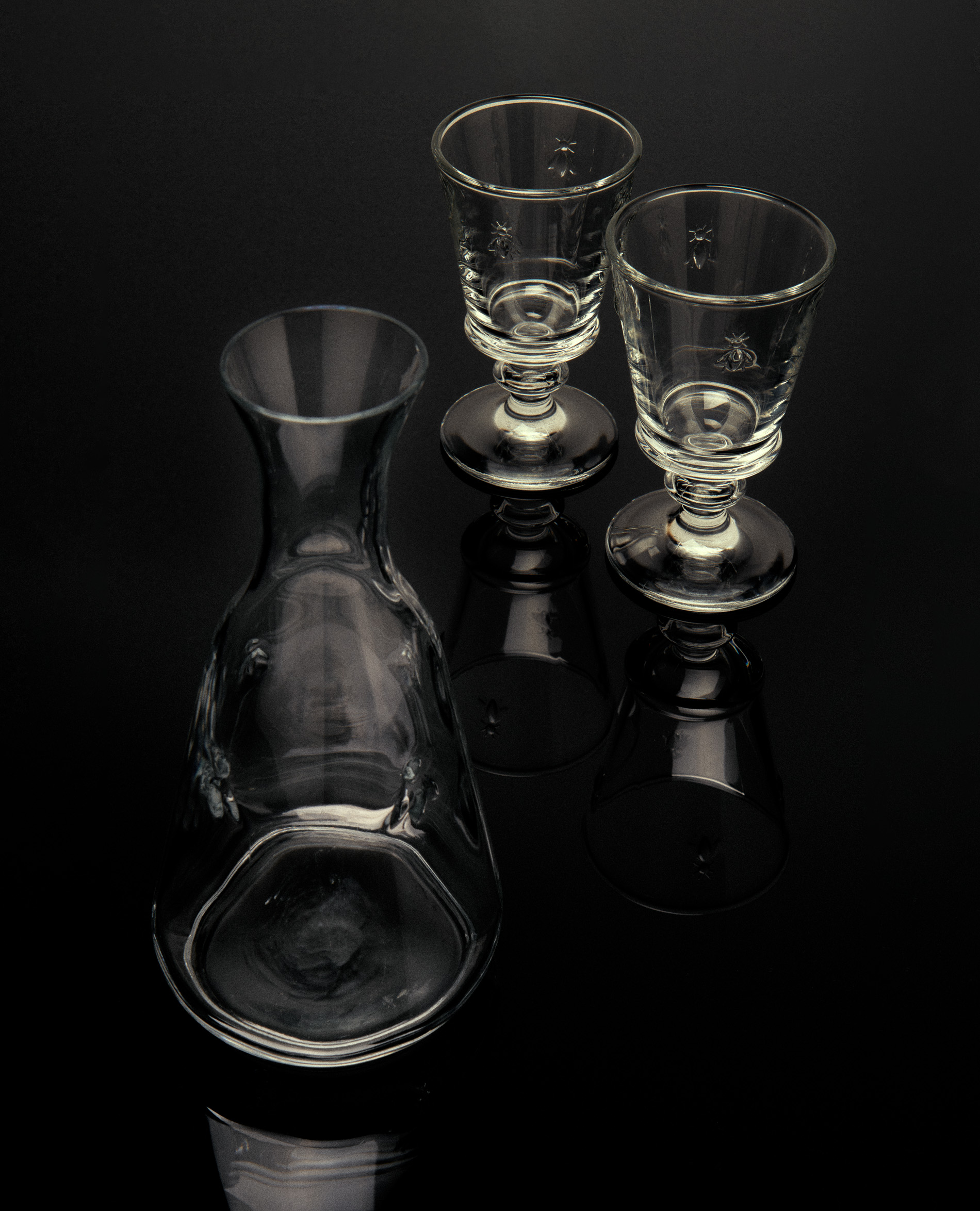 Still life of a clear glass carafe and two stemmed glasses on a glossy black surface with visible reflections in a minimalist composition.
