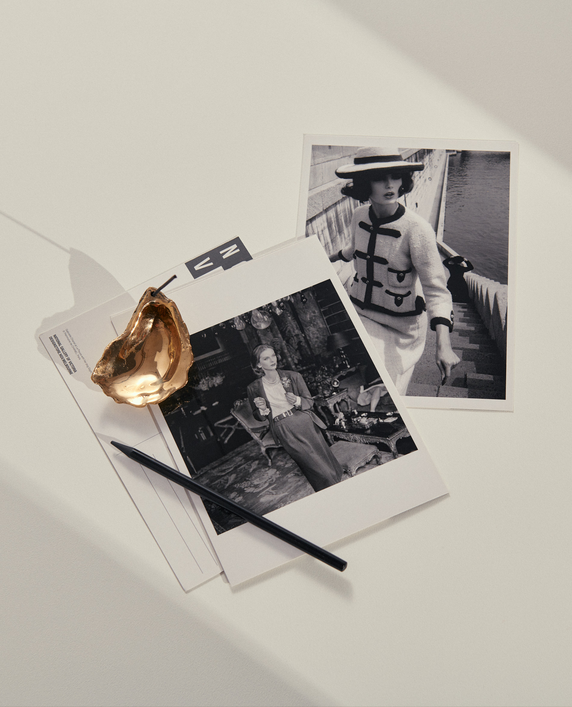 Black and white photographs of women in stylish outfits, a golden oyster shell, and a black pen are displayed on a white surface.