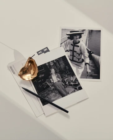 Black and white photographs of women in stylish outfits, a golden oyster shell, and a black pen are displayed on a white surface.