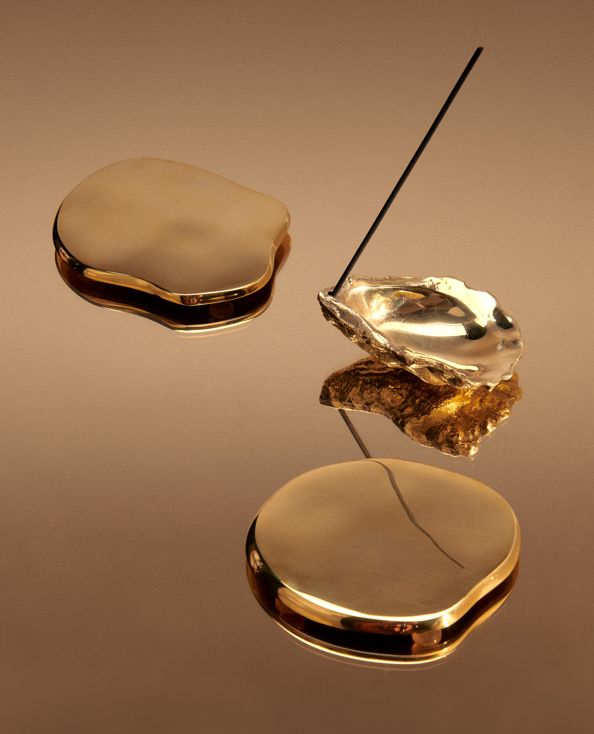 Still life of golden oyster shell incense holder with incense stick, and two decorative gold stone-shaped objects on a reflective surface.